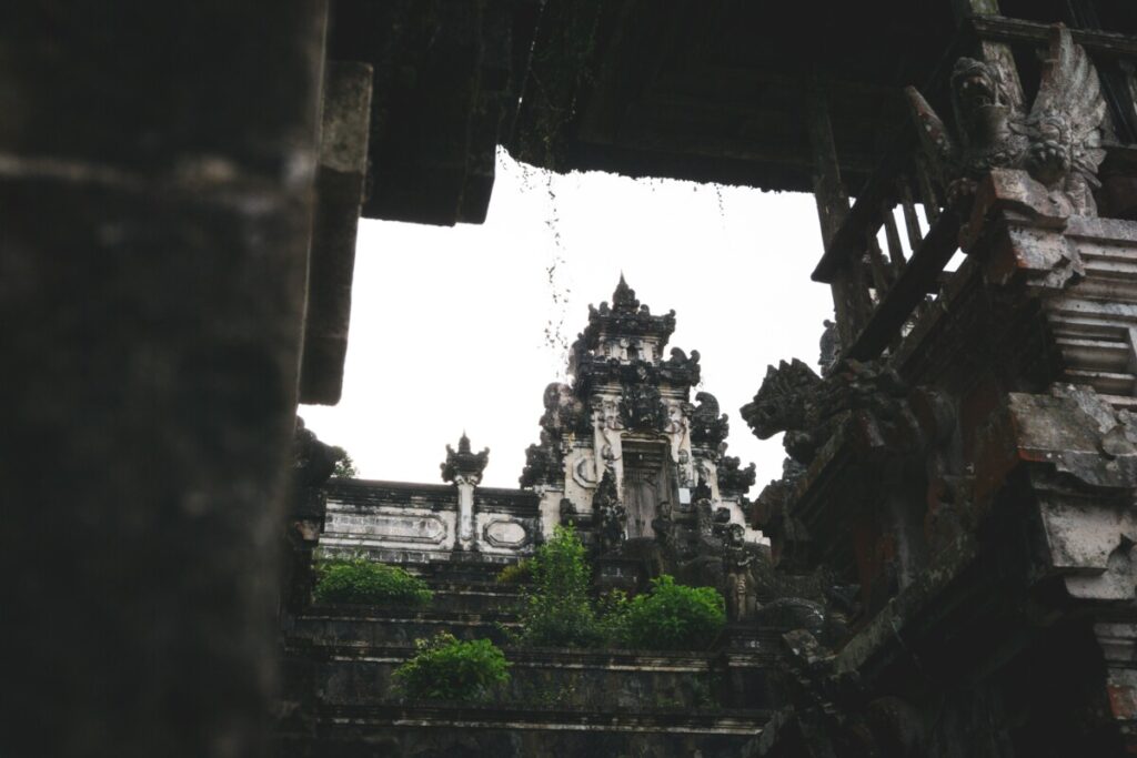 A moody, atmospheric image capturing the ancient, moss-covered stone gateways and statues of a Balinese temple, likely taken on an overcast day, which gives a sense of the mystical and historical essence of Bali's sacred architectural heritage.