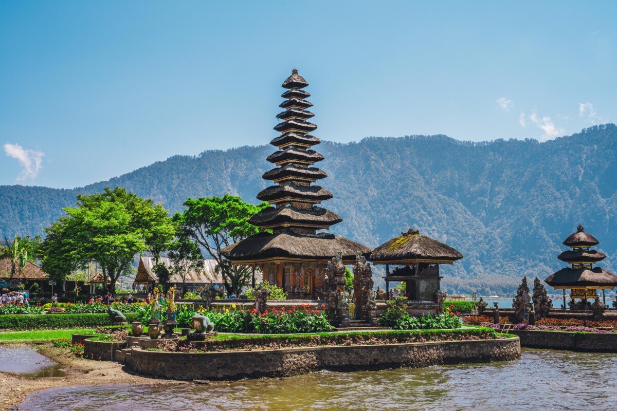 A vibrant photograph showcasing the iconic multi-tiered pagoda of Pura Ulun Danu Bratan Temple with a backdrop of mountains under a clear blue sky, reflecting the traditional Balinese architecture surrounded by lush greenery and vivid flowering plants at the temple's garden by the lakeside.
